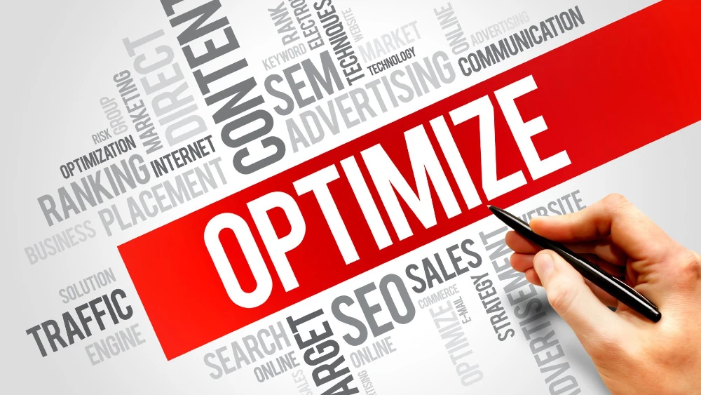 A conceptual image featuring the word “OPTIMIZE” in large red and white text across the center, surrounded by related digital marketing terms like SEO, content, traffic, placement, and advertising in varying shades of gray. A hand holding a pen is positioned in the lower right corner, implying action or planning.